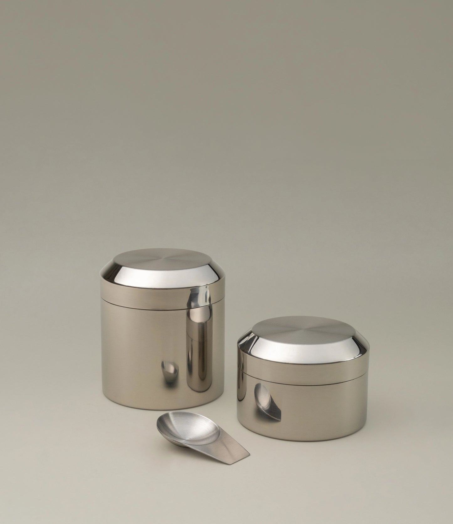 Sleek stainless steel tea canister set with a polished finish, featuring one tall and one shorter container. Both have rounded lids that seamlessly integrate with the cylindrical bodies, reflecting a minimalist design. A matching metallic scoop is placed in front, completing the set. The ensemble is set against a neutral background to emphasize its modern and elegant aesthetic.