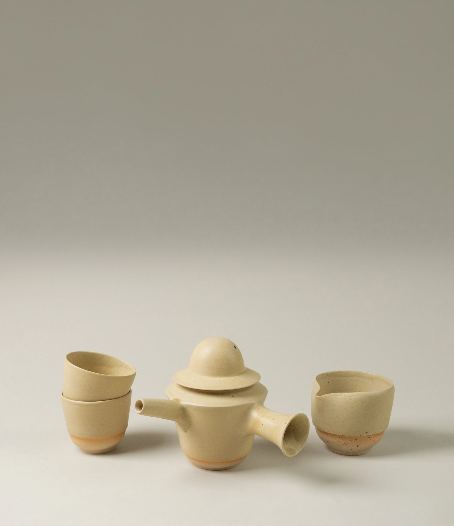 : "Omni Ceramics tea set displayed against a neutral background, featuring a clay teapot with a spout and lid, accompanied by two minimalist cups stacked on the left and one standalone pourer on the right.