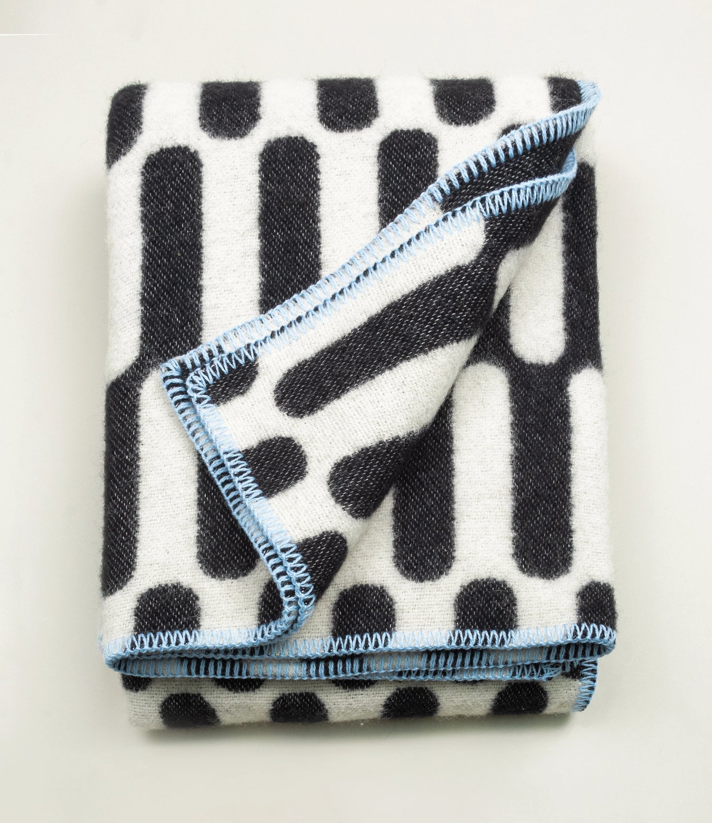 Throw Blanket From Tarta Gelatina coming in a black and white color. The pattern is similar to what you would see on op-art pictures. The edges are stiched with a light blue thread. In this picture the blanket is folded.